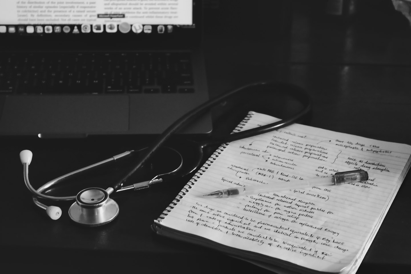 Stethoscope, notebook, vial, and laptop on a desk.